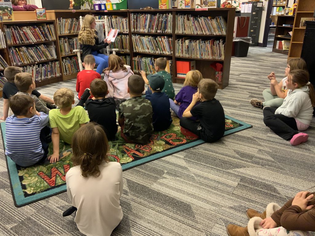 8th grader reads a book to elementary students who are sitting on the floor